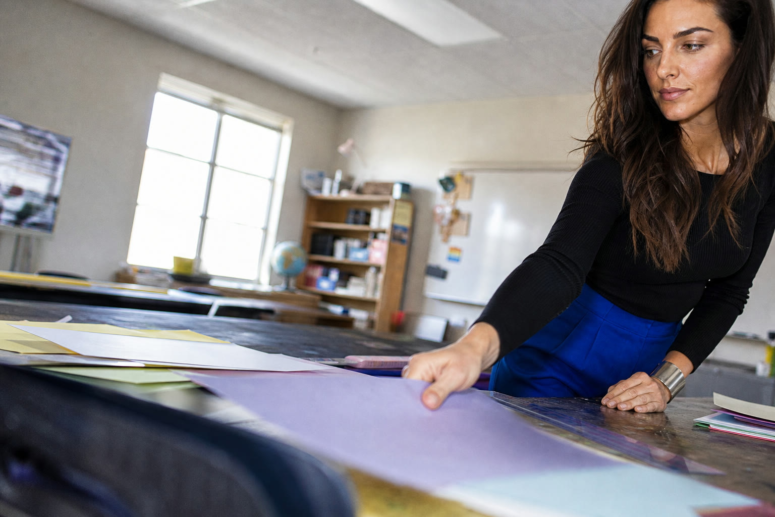 A self-possessed educator reaches across a classroom worktable in a vivid cobalt-and-black look.